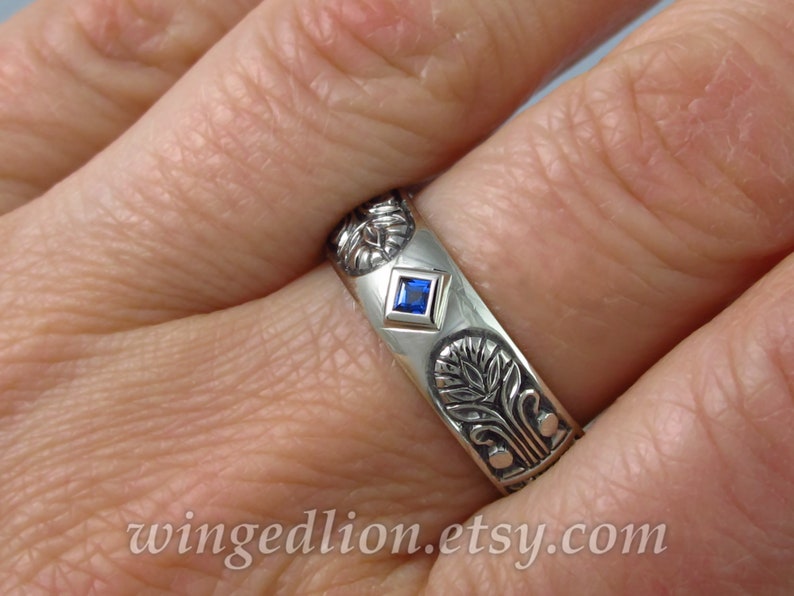 May include: A silver band ring with a blue gemstone set in a square bezel. The ring is decorated with an intricate floral design.