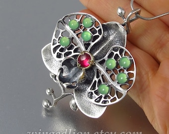 May include: A silver orchid pendant with green and red gemstones. The pendant is detailed with a delicate filigree design and hangs from a silver chain.