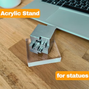 Pode incluir: Um suporte de acrílico transparente com um inserto de metal prateado em forma de quebra-cabeça. O suporte é projetado para exibir estátuas. O texto "Acrylic Stand" está no canto superior esquerdo da imagem. O texto "for statues" está no canto inferior direito da imagem.