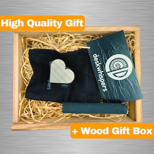 May include: A wooden gift box filled with brown packing material. The box contains a black pouch with a white heart-shaped object inside, a black tube, and a black card with the text "deskwhispers" and a logo.