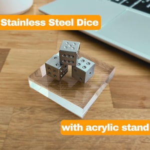 Può includere: Tre dadi in acciaio inossidabile con motivi a punti forati poggiano su un supporto in acrilico trasparente. I dadi sono argentati e il supporto ha un aspetto venato del legno. Il testo "Stainless Steel Dice" e "with acrylic stand" è visibile.