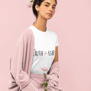 Puede incluir: Una camiseta blanca con el texto "FAITH > FEAR" en letras negras con un símbolo de corazón rosa. La modelo lleva un cárdigan rosa y una falda plisada rosa. La modelo tiene rosas rosas en el pelo.