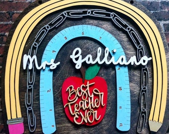 May include: A wooden wall sign featuring a layered rainbow design with yellow pencil arches, a light blue ruler arch, and a chain of paper clips. A red apple cutout in the centre displays the white text "Best Teacher Ever" and "Mrs Galliano".