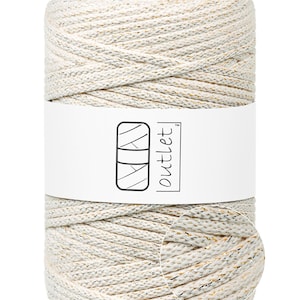 May include: A roll of off-white macrame cord with gold and silver flecks. A white band wraps around the centre, displaying a logo and the word "outlet". The cord is tightly wound, showcasing its textured appearance, ideal for crafting projects.