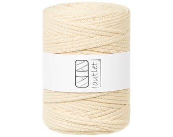 Puede incluir: Un carrete cilíndrico de cordón de macramé blanquecino. Una etiqueta blanca envuelve el cordón, mostrando un logotipo y la palabra "outlet". El cordón está bien enrollado, ideal para proyectos de manualidades.
