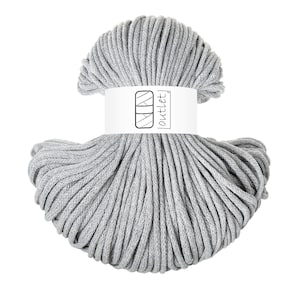 Peut inclure: Un écheveau de cordon gris clair présentant une texture tressée lisse. Une étiquette en papier blanc est enroulée autour du centre du cordon, affichant une icône graphique noire d'une pelote de laine et le texte "outlet" en police simple.