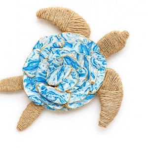 May include: A sea turtle wall decoration featuring four flippers and a head wrapped in natural tan jute twine. The shell is composed of layered, petal-like shapes painted with a marbled blue and white pattern, accented with gold-toned edges.