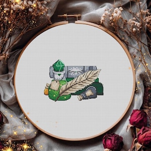 May include: A cross-stitch embroidery piece in a circular wooden hoop featuring a design of a green crystal, a scroll, a stack of books, and a large feather quill. The needlework uses various shades of green, brown, gray, and tan thread on white fabric.