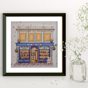 Pode incluir: Uma obra de arte emoldurada retrata uma ilustra&ccedil;&atilde;o detalhada de uma livraria. O edif&iacute;cio apresenta uma fachada azul com as palavras "BOOKS" e "CARDS" acima da entrada. A obra est&aacute; numa moldura preta, com fundo branco e um vaso de flores brancas.