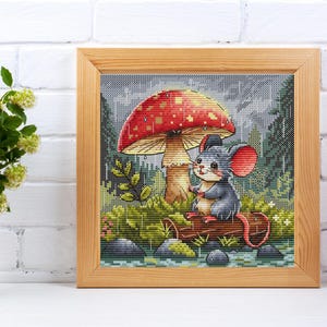 May include: A cute gray mouse sits under a red and white mushroom in a forest setting. The mushroom has white spots and a brown stem. The mouse is holding a small object in its paws. The background is a green and brown forest with rain falling.