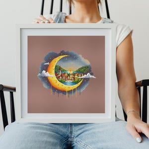 Può includere: Opera d'arte a punto croce incorniciata raffigurante una luna crescente con una scena di villaggio. La luna è gialla e arancione, con un cielo blu e nuvole. Il villaggio ha edifici con tetti rossi e una chiesa.
