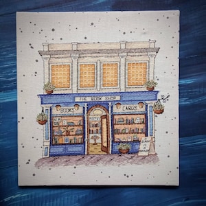 Pode incluir: Ilustra&ccedil;&atilde;o de uma livraria com um toldo azul que diz "The Book Shop". A loja apresenta livros, cart&otilde;es e cestos suspensos. Uma placa diz "A Book is a Dream". O edif&iacute;cio tem tr&ecirc;s janelas.
