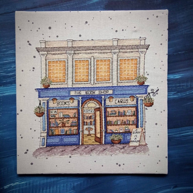 Pode incluir: Ilustra&ccedil;&atilde;o de uma livraria com um toldo azul que diz "The Book Shop". A loja apresenta livros, cart&otilde;es e cestos suspensos. Uma placa diz "A Book is a Dream". O edif&iacute;cio tem tr&ecirc;s janelas.
