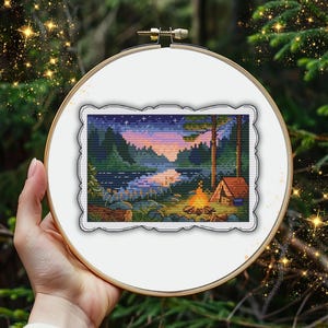 Può includere: Ricamo a punto croce di una scena di campeggio, con una tenda, un falò e un lago sotto un cielo stellato. L'opera è incorniciata in un telaio di legno, su uno sfondo sfocato di foresta.