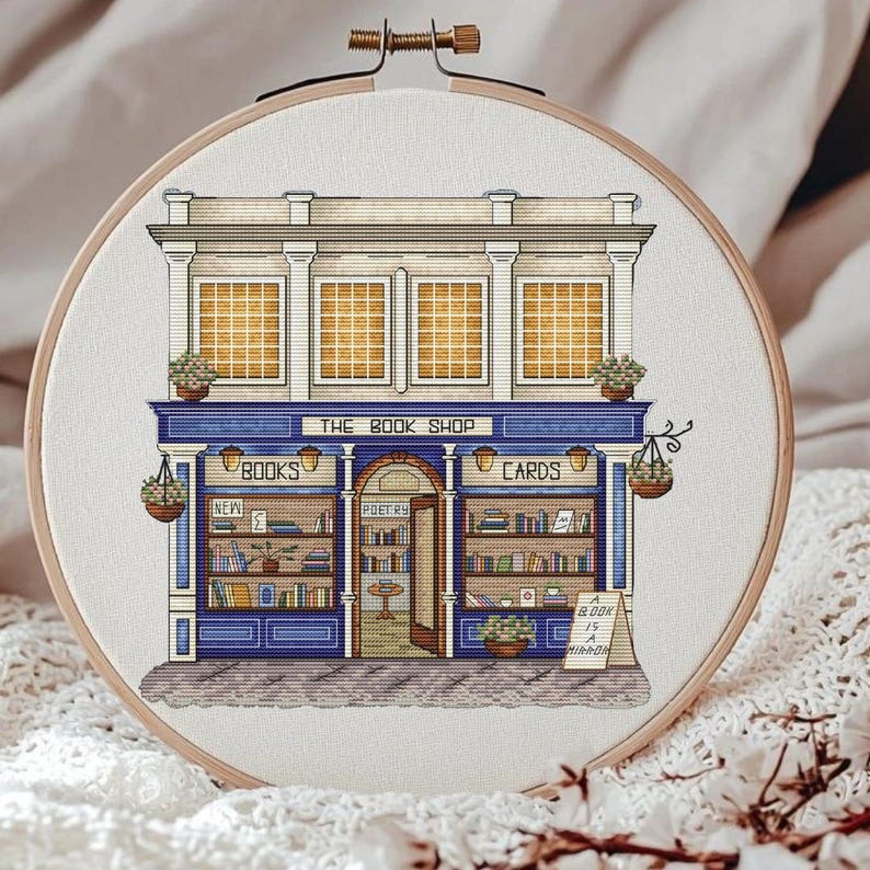 Pode incluir: Bordado em ponto cruz de uma livraria pitoresca. A loja apresenta um toldo azul com o texto "THE BOOK SHOP". A fachada inclui janelas, cestos de flores suspensos e as palavras "BOOKS" e "CARDS". Uma placa diz "A BOOK IS A MIRROR."