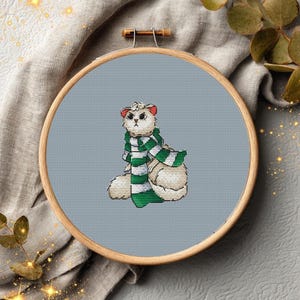 May include: Cross-stitch embroidery of a white cat wearing a green and white striped scarf. The cat is set against a light blue background within a wooden embroidery hoop. The design is detailed and whimsical, perfect for home decor.