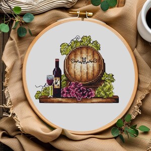 Könnte beinhalten: Kreuzstich-Stickerei in einem Holzrahmen mit einem Weinfass, einer Flasche, einem Weinglas und Trauben. Das Fass ist braun, die Trauben sind lila und grün, und der Wein ist rot. Die Stickerei befindet sich vor einem weißen Hintergrund.