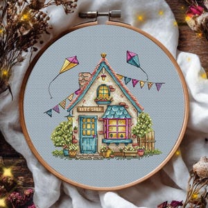 May include: A cross-stitch embroidery in a round wooden hoop featuring a house with a sign reading KITE SHOP. The design includes a blue door, a pink-framed window, colorful bunting flags, and two kites flying in the sky against a light blue fabric.