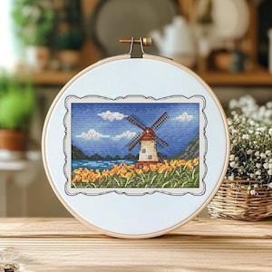 May include: A cross-stitch embroidery of a windmill scene, set in a wooden hoop. The artwork depicts a windmill with red and white details, set against a blue sky with clouds, a lake, and yellow flowers. The hoop is displayed on a wooden surface.