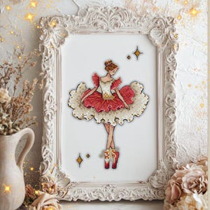 May include: A framed cross-stitch of a ballerina in a red and white tutu, pointe shoes, and a bun. The artwork is in a white ornate frame with a white background. Decorative gold stars are in the corners of the frame.