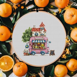 May include: A cross-stitch embroidery in a circular wooden hoop featuring a pink lemonade van with a house on its roof. The van has a blue and white striped awning, a sign reading LEMONADE, and a small chalkboard sign that says July 15.