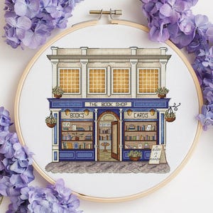 Pode incluir: Bordado em ponto cruz de uma livraria, emoldurado em um bastidor de madeira. A loja tem uma fachada azul e branca com o texto "THE BOOK SHOP". As janelas exibem livros e a placa diz "BOOKS" e "CARDS".