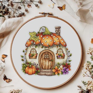 Puede incluir: Un diseño de punto de cruz de una casa calabaza caprichosa, dentro de un bastidor de bordado de madera. La casa presenta un techo de calabaza naranja con hojas verdes, una puerta de madera y detalles florales. El bastidor mide unos 20 cm de diámetro.