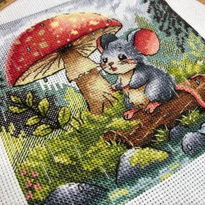 Może przedstawiać: Haft krzyżykowy przedstawiający szarą mysz siedzącą na pniu pod czerwono-białym grzybem. Scena rozgrywa się w lesie z zielonymi liśćmi, skałami i lekkim deszczem. Dzieło jest oprawione w drewnianą tamborkę.
