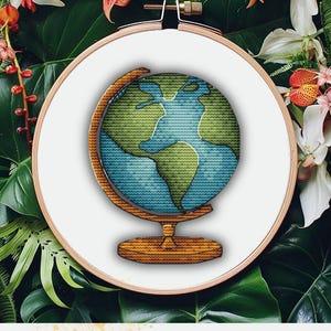 Op de afbeelding: Een borduurring met kruissteek met een wereldbolontwerp. De wereldbol is gestikt in blauw en groen garen, met de continenten en oceanen. De houten standaard en ring zorgen voor een natuurlijke uitstraling. De achtergrond is gevuld met weelderig groen gebladerte en bloemen.