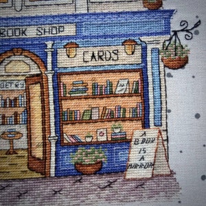 Pode incluir: Imagem em ponto cruz de uma livraria pitoresca com fachada azul e as palavras "BOOK SHOP" acima da entrada. A loja apresenta prateleiras de livros, um letreiro "CARDS" e um letreiro que diz "A BOOK IS A MIRROR".