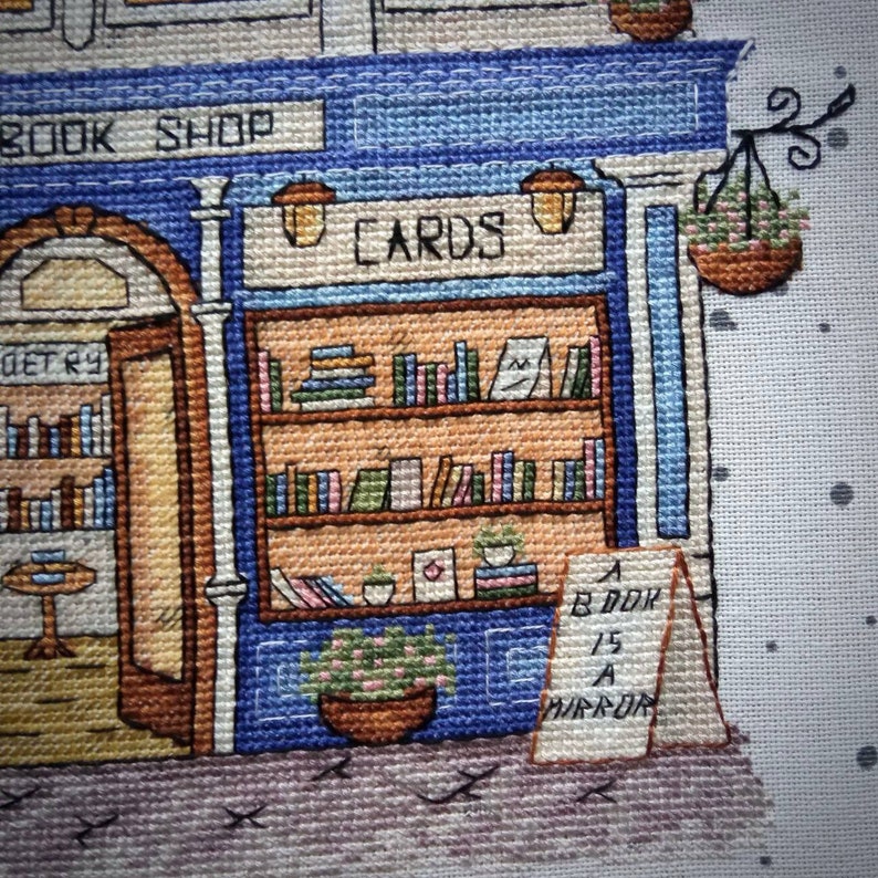 Pode incluir: Imagem em ponto cruz de uma livraria pitoresca com fachada azul e as palavras "BOOK SHOP" acima da entrada. A loja apresenta prateleiras de livros, um letreiro "CARDS" e um letreiro que diz "A BOOK IS A MIRROR".