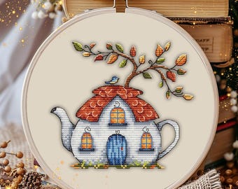 Op de afbeelding: Een kruissteekborduurwerk in een ronde houten borduurring met een witte theepot in de vorm van een huisje met een rood dak, een blauwe deur en gele ramen. Een kleine blauwe vogel zit op het dak onder een tak met oranje en groene bladeren.