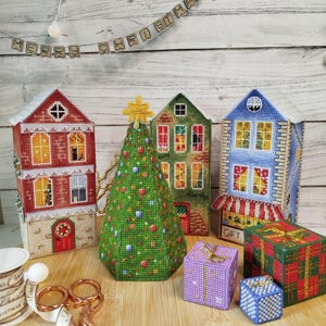 May include: A festive Christmas scene featuring handmade cross-stitch decorations. The display includes miniature houses, a Christmas tree, and wrapped gift boxes. A banner reads "MERRY CHRISTMAS".