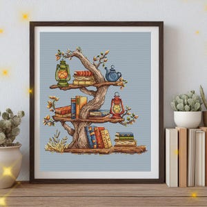 Puede incluir: Un bordado en punto de cruz enmarcado que muestra un árbol estilizado cuyas ramas sirven de estantes. El árbol sostiene pilas de libros coloridos, una tetera azul y dos linternas de estilo antiguo sobre un fondo azul claro.