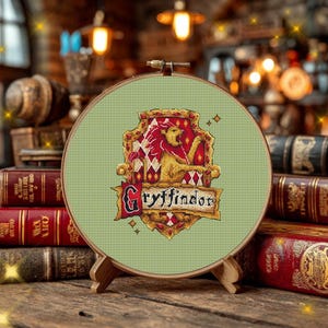 Puede incluir: Un bastidor de bordado circular de madera que contiene un diseño de punto de cruz de un león dorado sobre un escudo rojo y dorado. El texto Gryffindor está bordado en letras negras sobre una banda dorada en el centro, sobre tela verde claro.