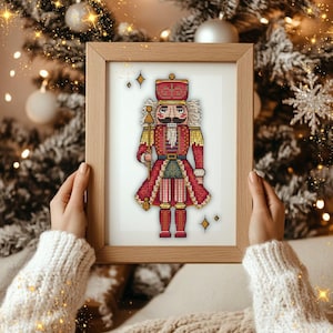 May include: A framed cross-stitch of a traditional Christmas nutcracker soldier in a wooden frame. The nutcracker is dressed in a red uniform with gold and green accents, holding a gold staff. The background is a blurred Christmas tree.