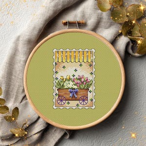 May include: A circular wooden embroidery hoop containing a cross-stitch pattern on light green fabric. The design features a brown cart filled with pink and yellow flowers, a blue bow, and a yellow striped awning, all framed by a white scalloped border.
