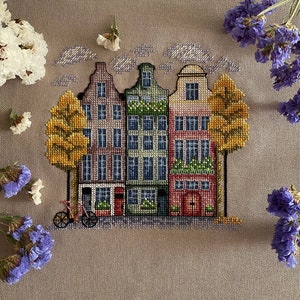 May include: Cross-stitch embroidery depicting three colourful houses with red doors and windows, set against a beige background with yellow trees and a bicycle in front.