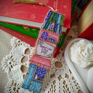 Op de afbeelding: Een kleurrijke, met kralen bezette boekenlegger in de vorm van huizen. Het heeft een blauwe deur, roze muren en een groen dak. De tekst "Tomorrow a new page" is zichtbaar. Een gouden kwast hangt eraan. De boekenlegger ligt op een kanten kleedje.