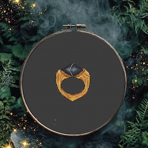May include: A circular wooden embroidery hoop containing a dark gray fabric background. Centered on the fabric is a cross-stitch design of a gold ring featuring a large, dark, faceted gemstone at the top, rendered with detailed golden thread.