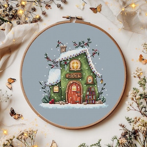 May include: Cross-stitch embroidery of a whimsical green house with a red door and the word "BOOKS" above it. The house is set in a snowy scene, framed in a wooden hoop. Butterflies and floral accents surround the hoop.