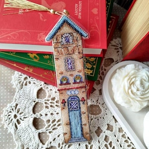 以下が含まれることがあります： ベージュ、青、茶色のクロスステッチ刺繍が施された、家の形をした手作りのブックマーク。青い屋根、青いドア、金のタッセルが付いています。赤い本の手前に置かれています。