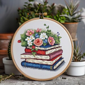 May include: Cross-stitch embroidery in a wooden hoop. The design features a stack of books with a colorful floral arrangement on top. The flowers include pink, white, and blue blooms with green foliage. The books are red, pink, and blue.