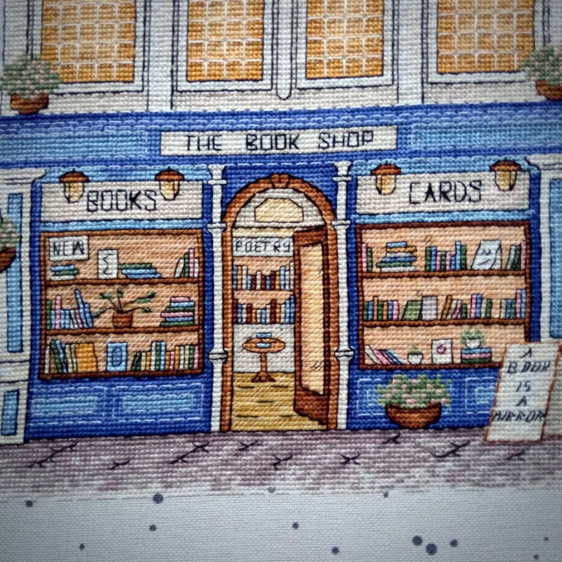 Pode incluir: Uma representa&ccedil;&atilde;o em ponto cruz de uma fachada de livraria pitoresca. A loja, pintada em azul e branco, apresenta o texto "THE BOOK SHOP" acima da entrada. Prateleiras cheias de livros e cart&otilde;es, com a palavra "POETRY" acima da porta aberta.