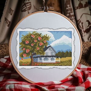 May include: A circular wooden embroidery hoop containing a cross-stitch pattern of a house with a porch, a large tree with red apples, a yellow field, and a blue sky with white clouds, all framed by a decorative white border on light fabric.