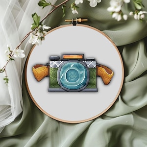 May include: A cross-stitch embroidery design of a camera centred in a round wooden hoop. The camera features a large blue lens, a green body with a textured pattern, and orange side grips. The top of the camera has a rectangular orange accent.