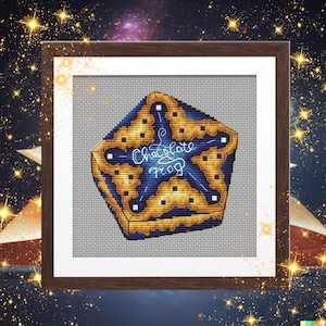 Puede incluir: Arte de punto de cruz enmarcado con un diseño de rana de chocolate. La obra de arte muestra una forma geométrica azul y dorada con las palabras "Chocolate Frog" en cursiva. El fondo es una tela gris claro.