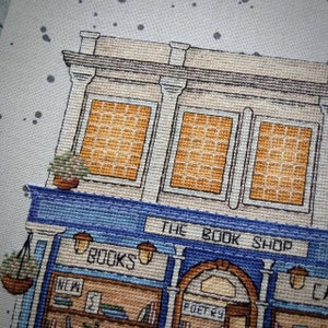 Pode incluir: Representa&ccedil;&atilde;o em ponto cruz de uma fachada de livraria. O edif&iacute;cio apresenta um toldo azul com o texto "THE BOOK SHOP". As janelas t&ecirc;m um padr&atilde;o de grade, e a loja tem cestos de flores suspensos e uma placa que diz "BOOKS".