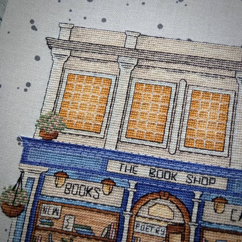 Pode incluir: Representa&ccedil;&atilde;o em ponto cruz de uma fachada de livraria. O edif&iacute;cio apresenta um toldo azul com o texto "THE BOOK SHOP". As janelas t&ecirc;m um padr&atilde;o de grade, e a loja tem cestos de flores suspensos e uma placa que diz "BOOKS".