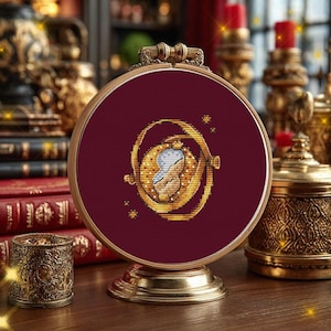 Puede incluir: Un bastidor de bordado circular que contiene un diseño de punto de cruz de un colgante giratiempo dorado. El colgante presenta un reloj de arena central con arena azul claro, rodeado de anillos giratorios sobre un fondo de tela burdeos.